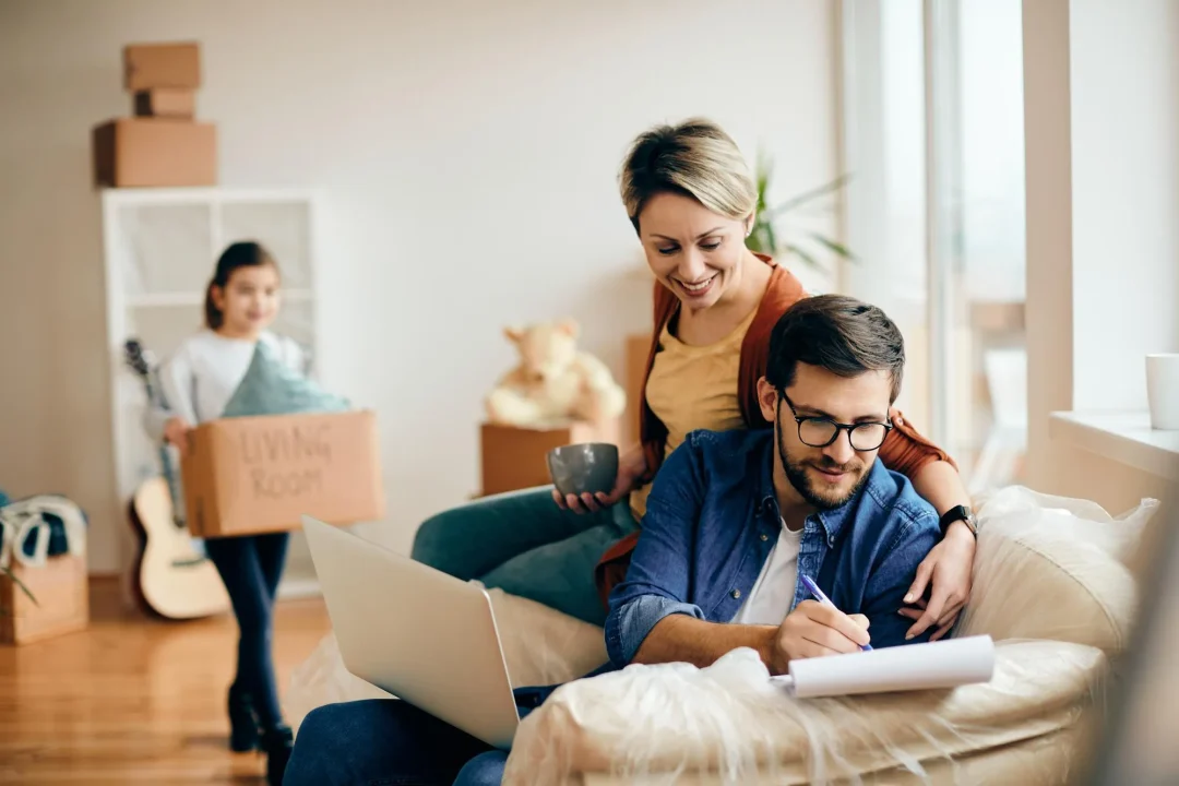 Familie in ihrer neuen Wohnung, Vater am Laptop, Mutter und Kind im Hintergrund mit Kartons