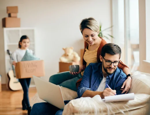 Familie in ihrer neuen Wohnung, Vater am Laptop, Mutter und Kind im Hintergrund mit Kartons