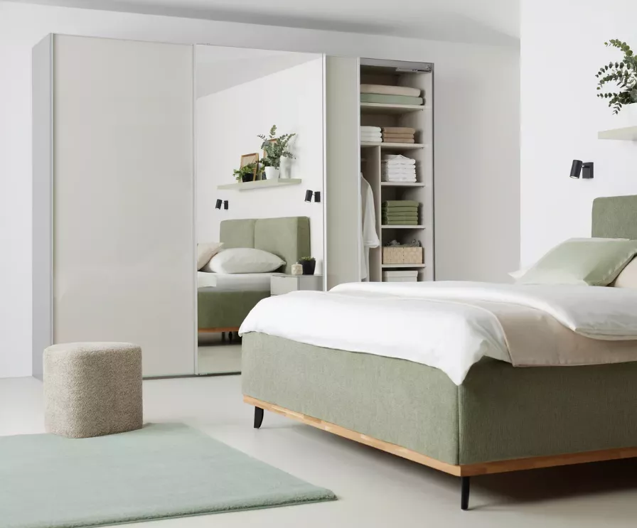 Hochglanzmöbel Trends 2025 – modernes Schlafzimmer mit weißem Hochglanz-Kleiderschrank, Spiegeltüren, grünem Boxspringbett und minimalistischer Dekoration.