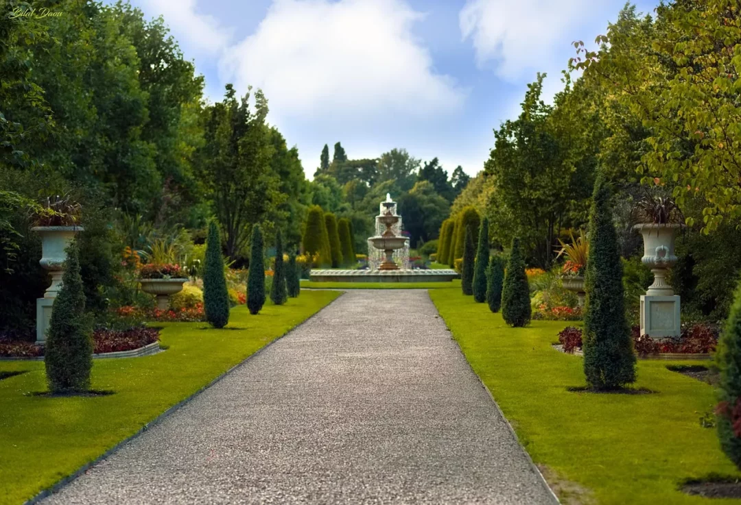 Garten gestalten mit breitem Kiesweg, geschnittenen Koniferen, dekorativen Pflanzgefäßen, farbenfrohen Beeten und großem Brunnen im Hintergrund.