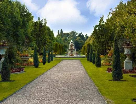 Garten gestalten mit breitem Kiesweg, geschnittenen Koniferen, dekorativen Pflanzgefäßen, farbenfrohen Beeten und großem Brunnen im Hintergrund.
