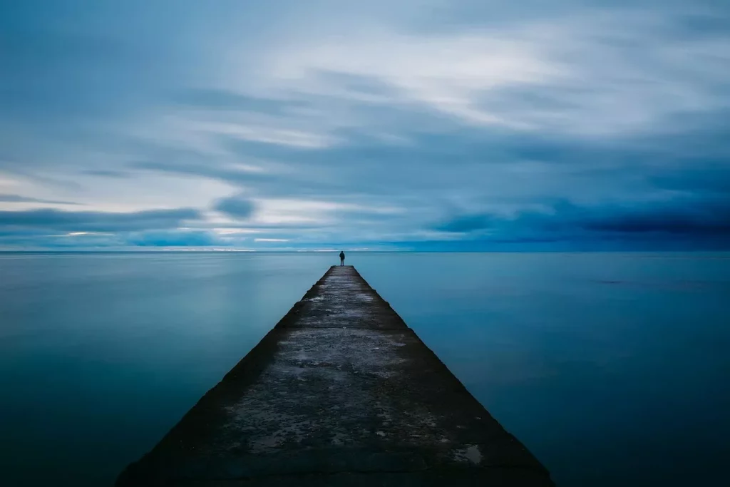 Eine Person steht allein auf einem langen Steg, der in ein ruhiges, weites Meer führt, unter einem blauen Himmel – Symbol für Minimalismus und Klarheit.