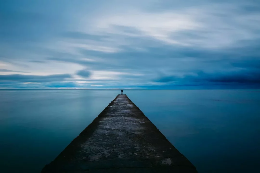 Eine Person steht allein auf einem langen Steg, der in ein ruhiges, weites Meer führt, unter einem blauen Himmel – Symbol für Minimalismus und Klarheit.