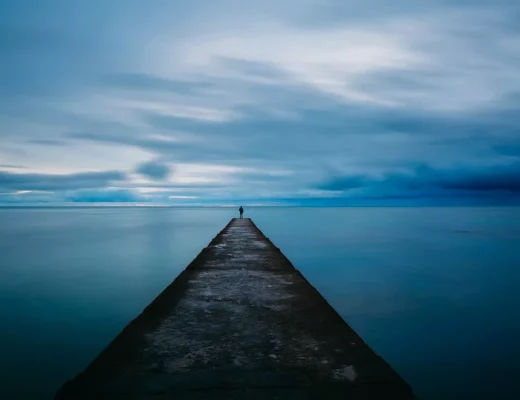 Eine Person steht allein auf einem langen Steg, der in ein ruhiges, weites Meer führt, unter einem blauen Himmel – Symbol für Minimalismus und Klarheit.