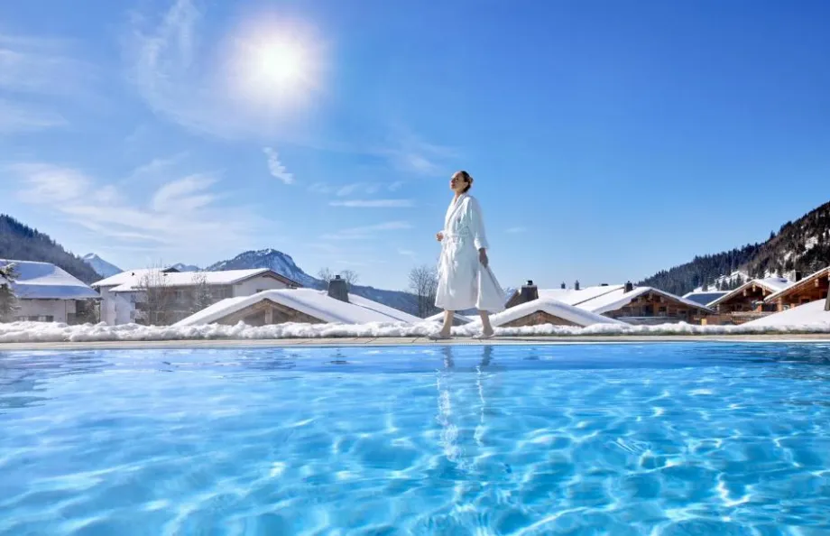 Frau im Bademantel am beheizten Infinity-Pool des Panoramahotels Oberjoch vor schneebedeckten Chalets und Alpenkulisse