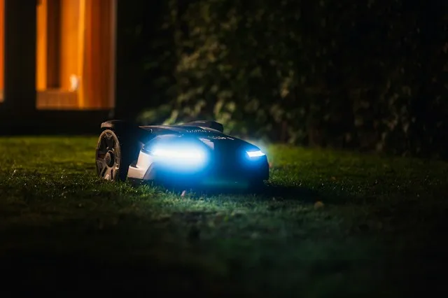 Mähroboter mit eingeschalteten LED-Scheinwerfern fährt nachts über eine Rasenfläche im Garten.
